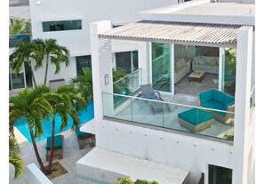 Um edifício moderno com varanda de vidro que dá para uma área de piscina, mobiliada com assentos externos e palmeiras.