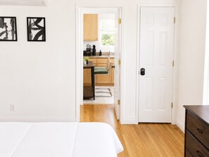 Een slaapkamer met een bed, een houten dressoir en een witte deur die leidt naar een keuken met houten kastjes en een raam.