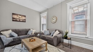 Ein Wohnzimmer mit zwei grauen Sofas, einem Holztisch, einer Uhr und einem Fenster mit Jalousien.