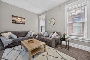 Uma sala de estar com dois sofás cinza, uma mesa de centro de madeira, um relógio e uma janela com persianas.
