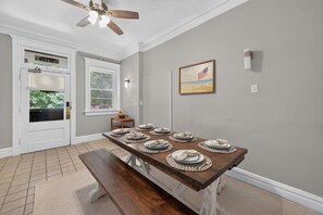 Uma sala de jantar com uma mesa comprida posta para seis pessoas, um ventilador de teto, uma porta com persianas e um quadro na parede.