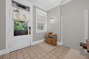 Um corredor com uma porta branca, uma janela com persianas, um banco de madeira e um candeeiro fixado na parede.