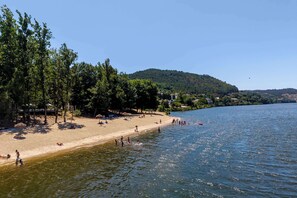 Ein Strand mit badenden und sonnenden Personen, umgeben von Bäumen und Hügeln.