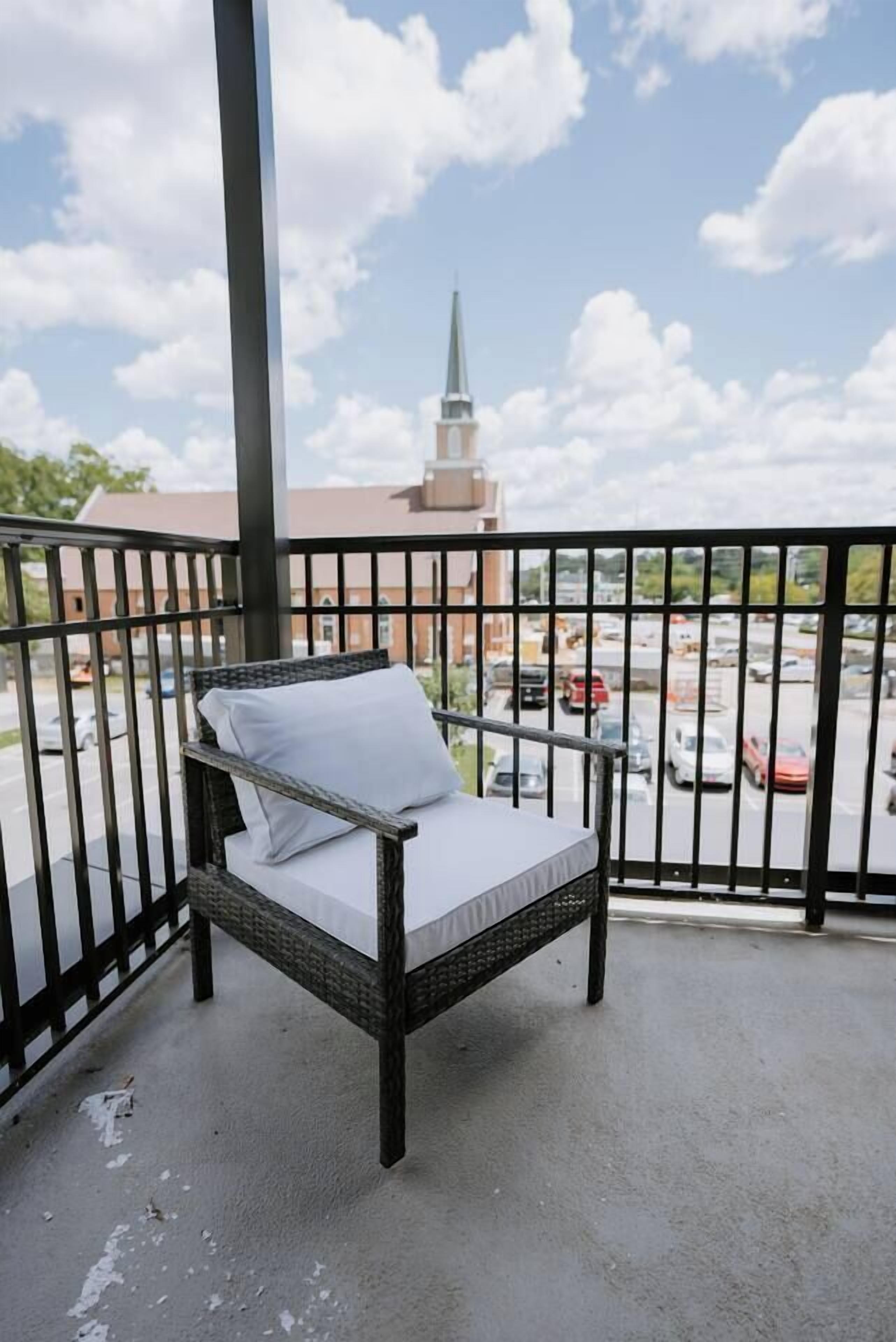 A balcony with a wicker chair and a view of a parking lot and a building with a spire.