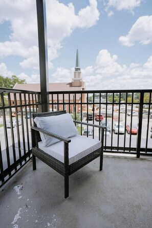 A balcony with a wicker chair and a view of a parking lot and a building with a spire. - Lovely 2BR · Williams-Brice & Fort Jackson (Columbia)