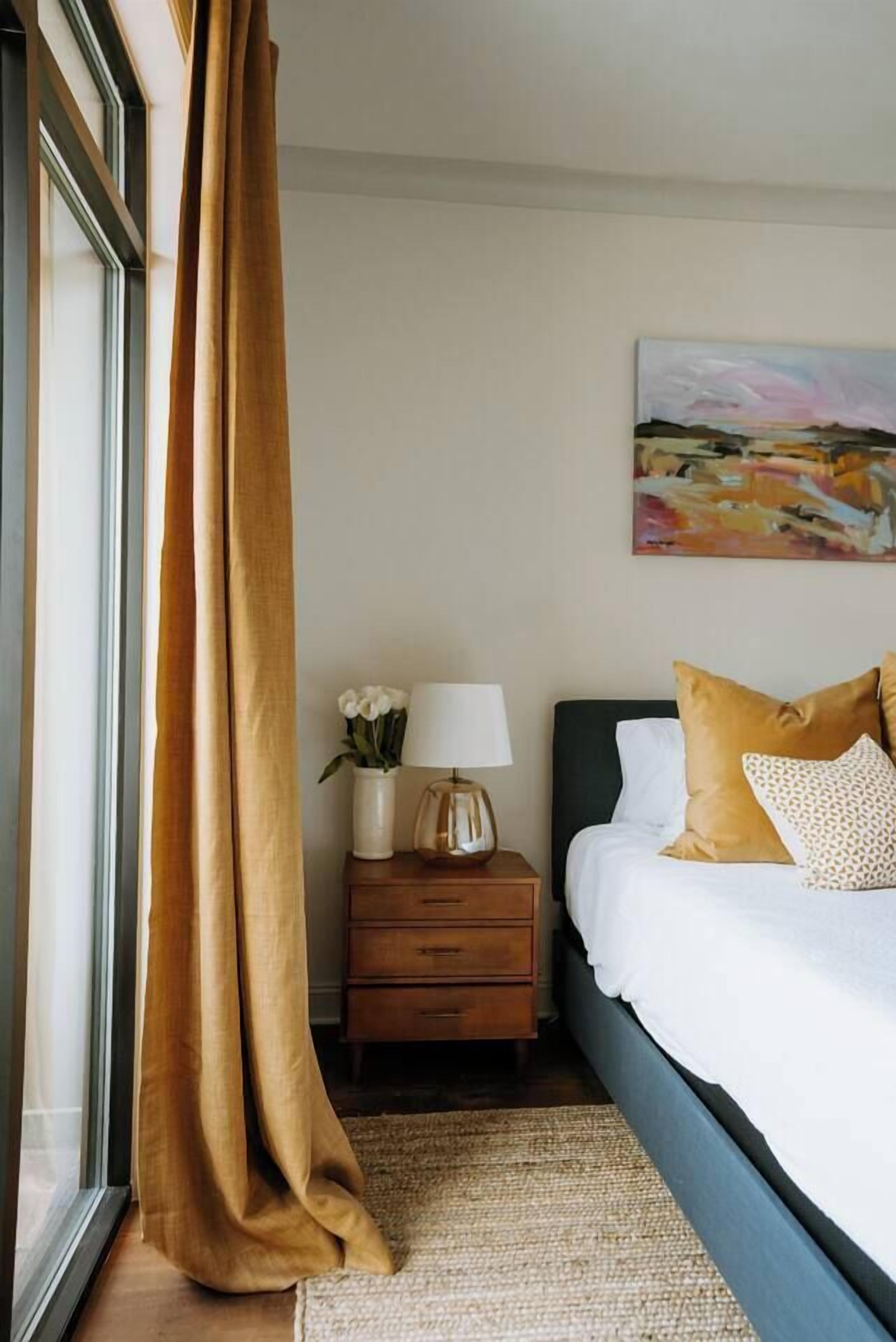 Un dormitorio con cama, mesita de noche, lámpara, un jarrón con flores y un cuadro en la pared.