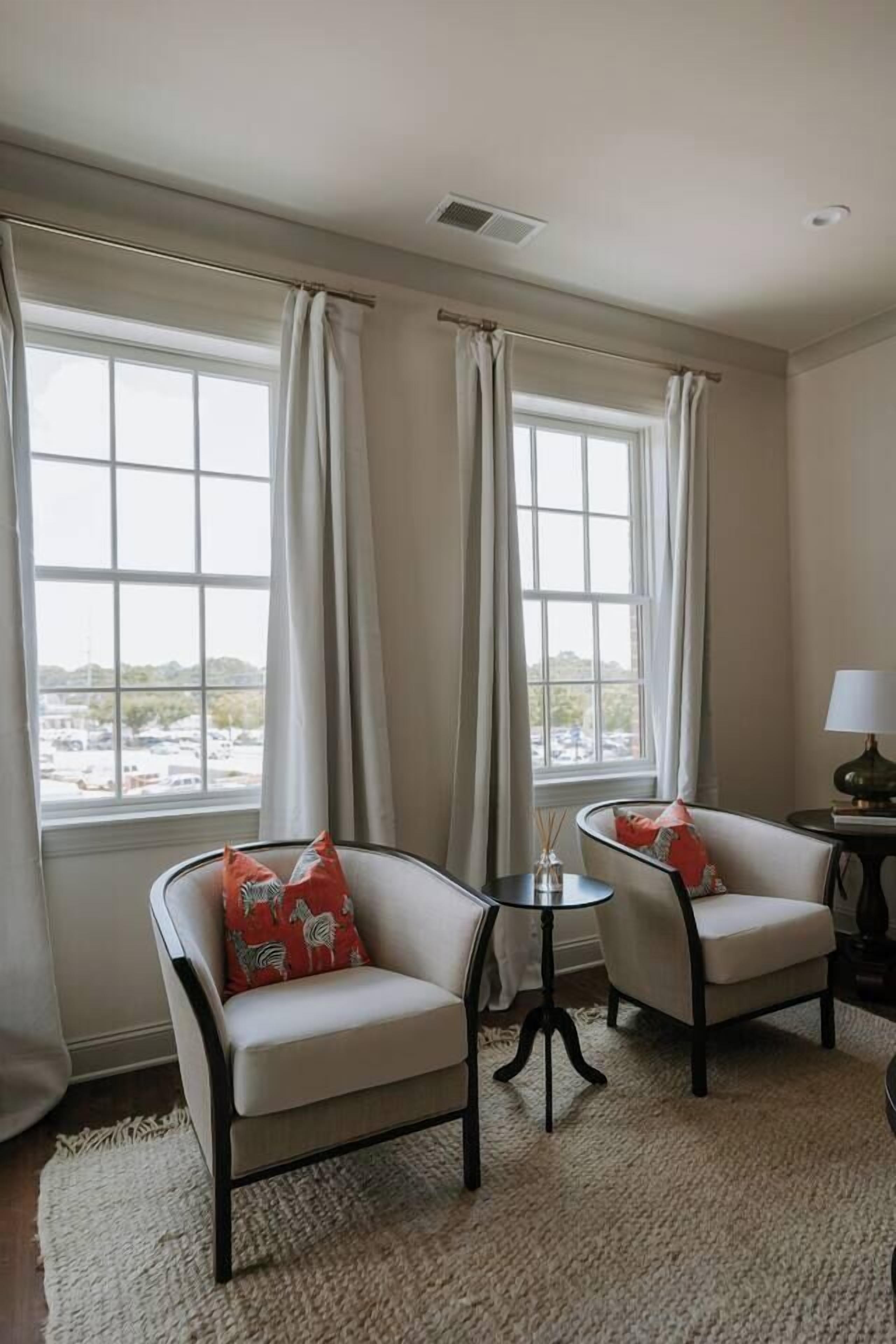 A room with two armchairs, a small table, and a view of a parking lot through large windows.