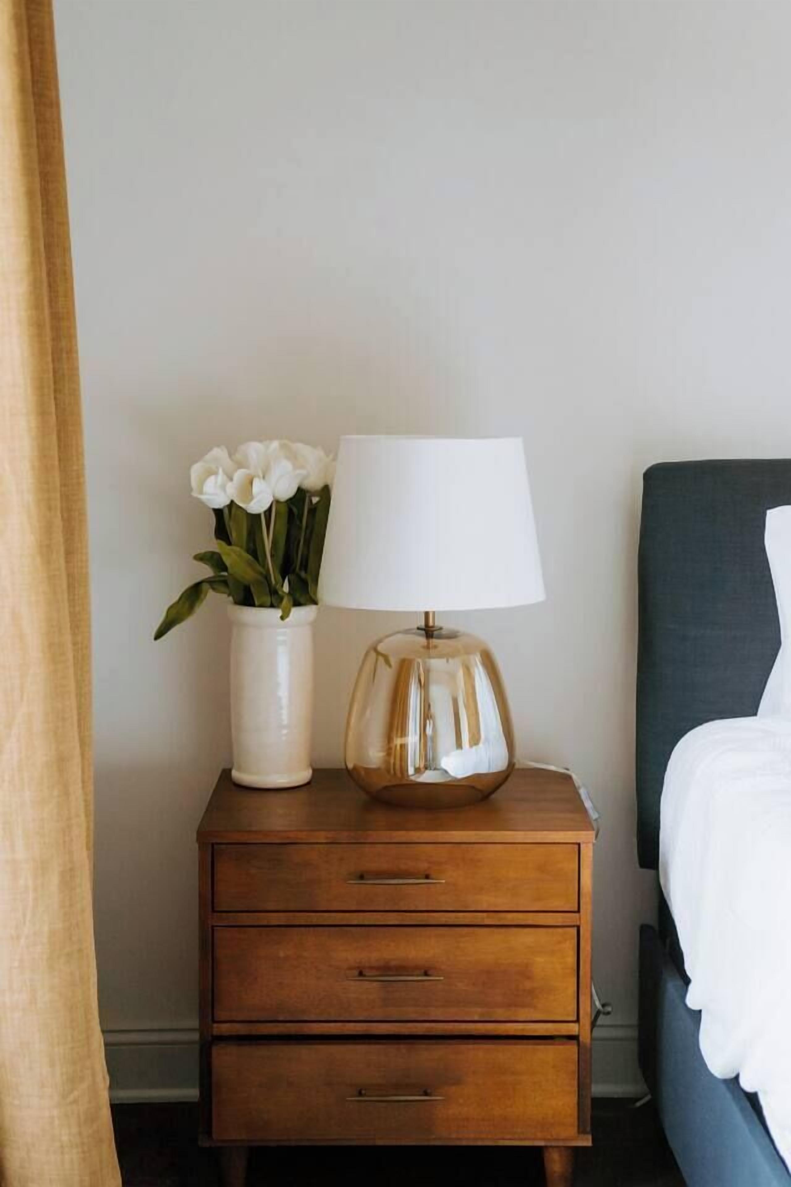 Een houten nachtkastje met een witte vaas vol bloemen en een lamp.