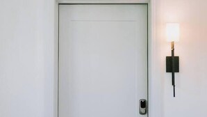 Eine weiße Tür mit Schlüsselloch und Klingel, eine Wandleuchte sowie ein Teppichboden.