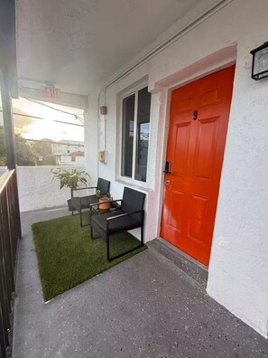 A balcony with a red door, a window, a small table with two chairs, and a potted plant.