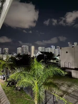 A nighttime view of a cityscape with high-rise buildings, a palm tree, and a paved area with parked motorcycles.