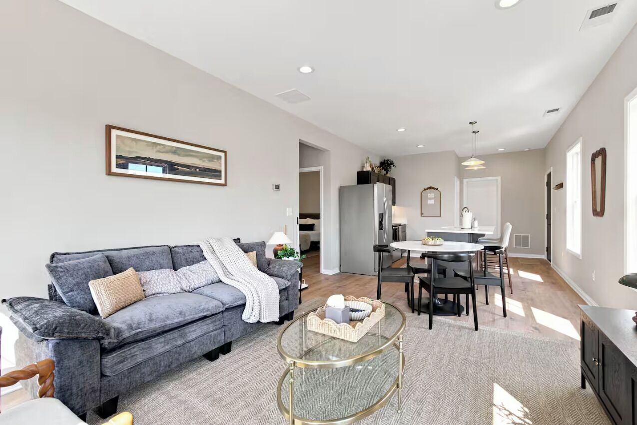 A modern living room with a grey sofa, a glass coffee table, and a dining area with a white table and chairs.