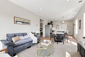 A modern living room with a grey sofa, a glass coffee table, and a dining area with a white table and chairs. - Stylish Chevy Chase Stay - Walk to Dining and UK (Lexington)