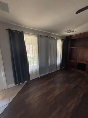 A room with a large window, dark curtains, a wooden floor, and a ceiling fan.