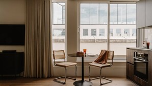 A modern living room with a flat-screen TV, a round glass table, two wicker chairs, a large window with a view of buildings, and a kitchen area with a dark countertop and cabinets. - Fulham Residences Seven (London)