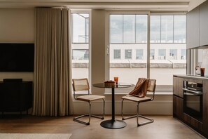 Ein modernes Wohnzimmer mit Flachbildfernseher, einem runden Glastisch, zwei Korbstühlen, einem großen Fenster mit Blick auf Gebäude und einer Küchenzeile mit dunkler Arbeitsplatte und Schränken.