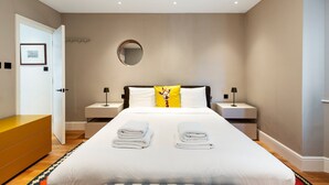 Ein modernes Hotelzimmer mit einem großen Bett, zwei Nachttischen, einem runden Spiegel und einer Tür zu einem anderen Raum.