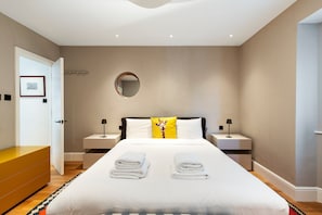 A modern hotel room with a large bed, two bedside tables, a round mirror, and a door leading to another room. - Chiswick Four-Bed Garden Retreat (London)