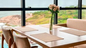 A dining table with placemats, a vase with flowers, and a view of a rocky coastline through large windows. - Blue Sea Hotel Luxury Suite Oceanfront Itapema SC (Itapema)