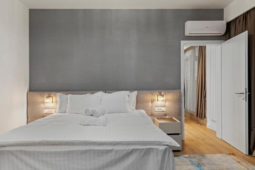 A hotel room with a bed, bedside tables, a wall-mounted air conditioner, and a door leading to a hallway.