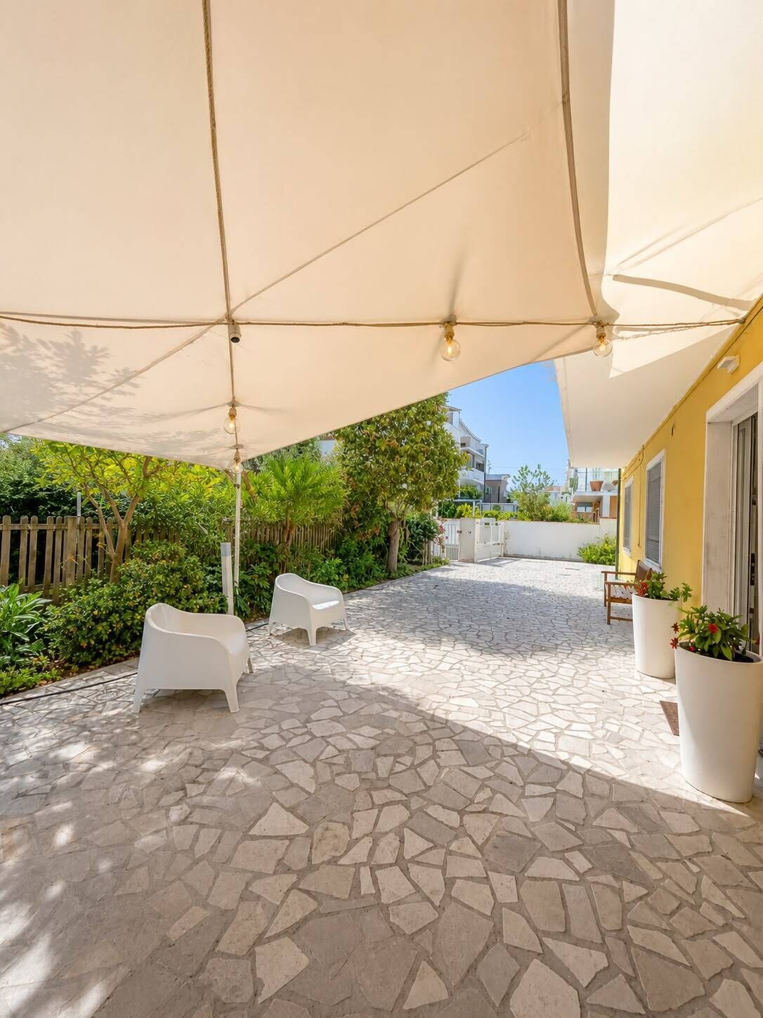 An outdoor patio area with stone flooring, white chairs, and a large canopy providing shade.