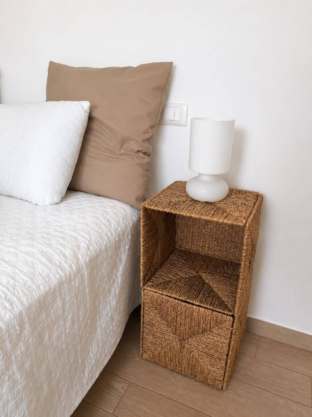 A bed with a white quilt and two pillows, a white bedside lamp on a woven nightstand, and a power outlet on the wall.