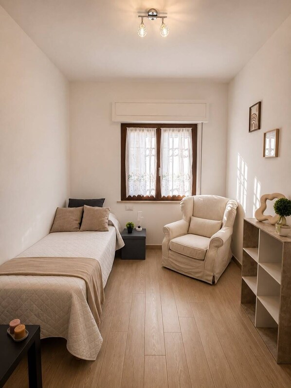 A minimalist bedroom with two beds, a chair, a nightstand, and a window with curtains.