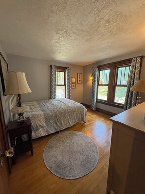 A bedroom with a bed, bedside tables, a round rug, and two windows with curtains.