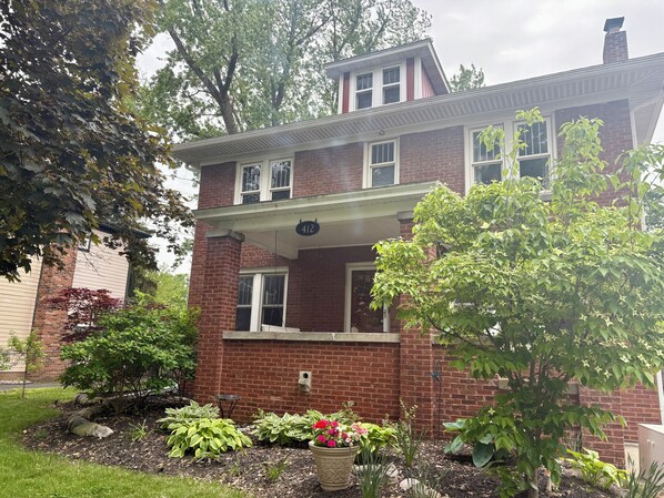 A two-story brick house with a covered front porch, a mailbox, and a garden with various plants and flowers.