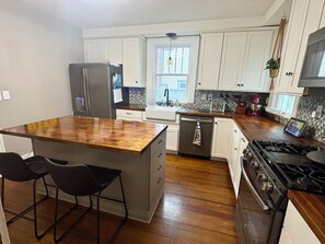 Una cocina moderna con armarios blancos, una isla de madera con taburetes, un refrigerador de acero inoxidable, un lavavajillas, una estufa a gas y un ventanal que proporciona luz natural.