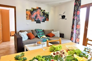 A living room with a sofa, a dining table set with food and drinks, and a wall with abstract art. - Bošković-Apartment-2, Modern Family Apartment with Balconies | 2 Bedrooms 6 pers (Rudina)