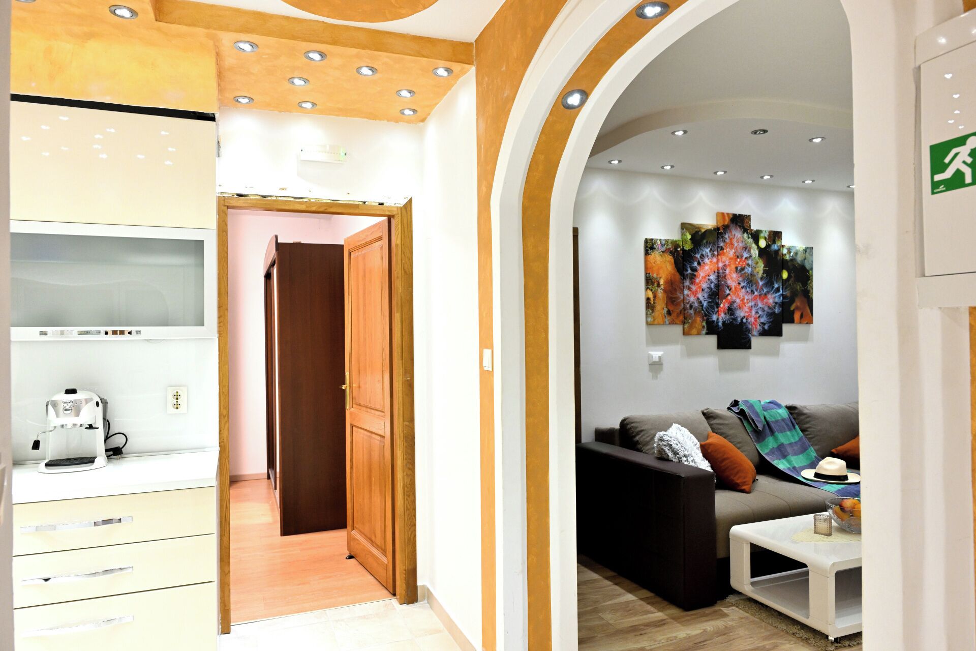 Ein modernes Interieur mit einer Holztür, die zu einem Flur führt, einem Wohnbereich mit Sofa und Kunstwerken sowie einer Küchenzeile mit einer Kaffeemaschine.