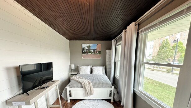 A bedroom with a bed, a TV, a window with curtains, and a view of the outside.