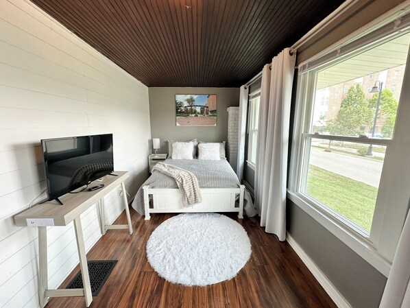 A bedroom with a bed, a TV, a window with curtains, and a view of the outside.