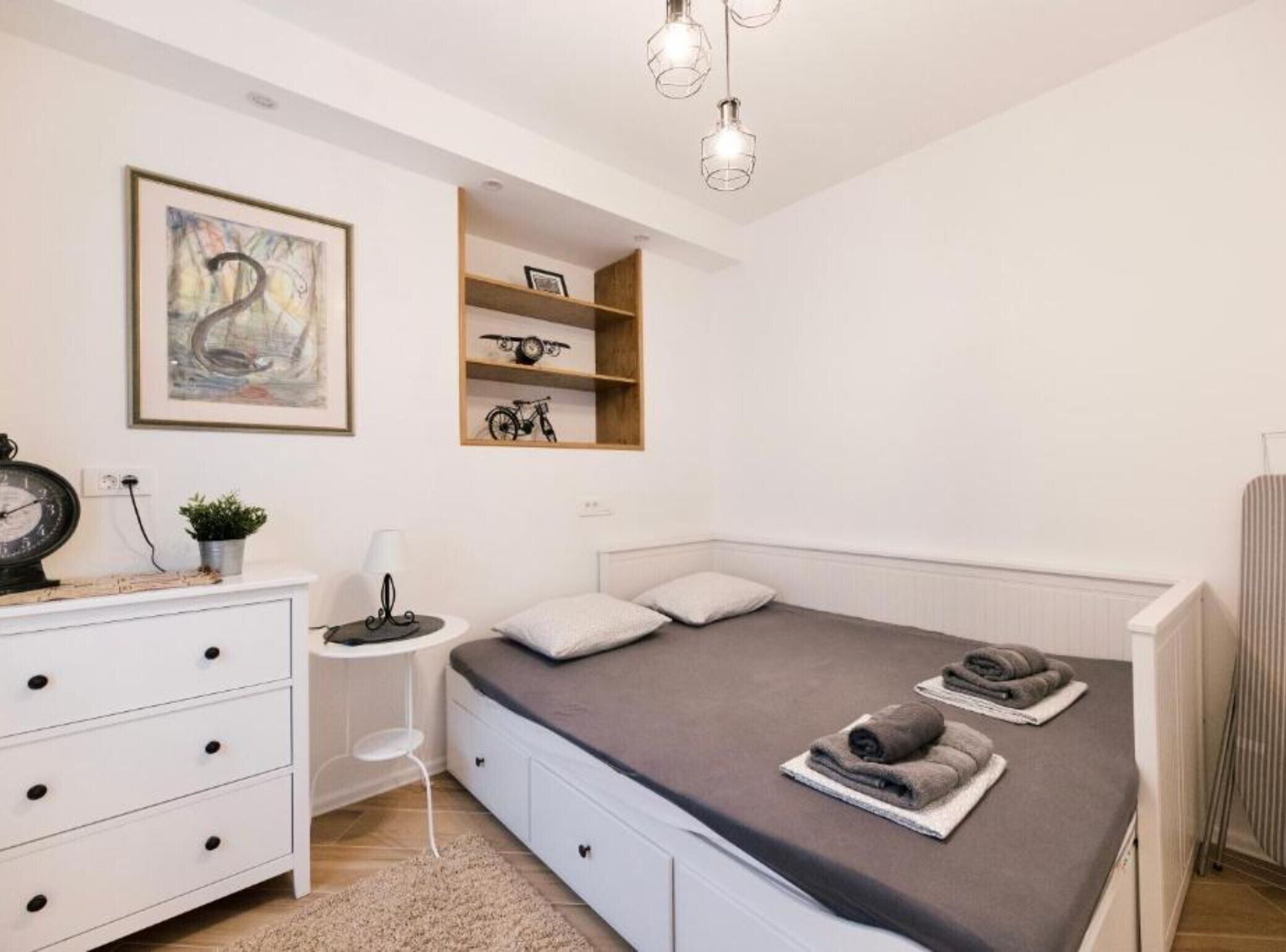 A bedroom with a bed, a dresser, a small table, a chair, a shelf with items, and a painting on the wall.