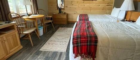 A bedroom with a wooden floor, a bed with a plaid blanket, a desk with a chair, a dresser, and a window with curtains.
