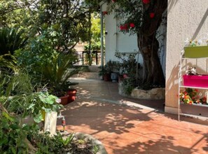 A garden pathway leading to a house with potted plants and a tree with red flowers. - Two Bedroom Apartment with Sea View and Balcony (Dubrovnik)