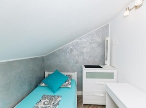 Ein Schlafzimmer mit schrägem Dach, einem Bett mit gemusterten Betttextilien, einem Nachttisch, einem Schreibtisch mit Stuhl und einer Wandleuchte.