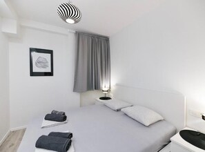 Ett minimalistiskt hotellrum med en säng, ett sängbord, en lampa och en inramad tavla på väggen.