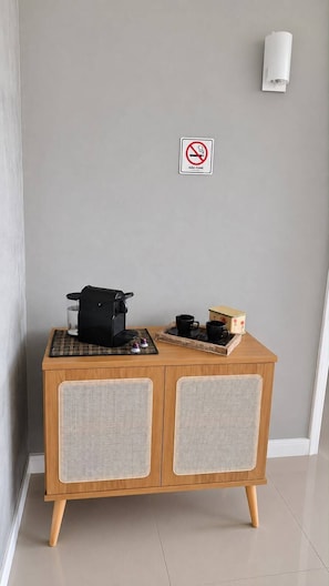 Ein Holzkabinett mit gewobenen Paneelen, eine Kaffeemaschine und zwei Tassen auf einer Ablageplatte, vor einer grauen Wand mit einem Rauchverbot-Schild.