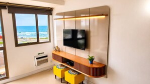 Un saló modern amb televisió de pantalla plana penjada a la paret, una taula de consola de fusta, dos otomans grocs i vista a l'oceà a través d'una finestra gran.