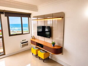 Ein modernes Wohnzimmer mit einem Wand-Fernseher, einem Holzkonsolentisch, zwei gelben Poufs und Blick auf den Ozean durch ein großes Fenster.