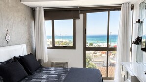 Un dormitori amb un llit, una finestra gran amb vista a la platja i un prestatge blanc.
