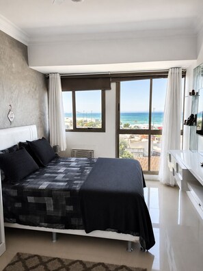 Ein Schlafzimmer mit einem Bett, einem großen Fenster mit Blick auf den Strand und einer weißen Kommode.