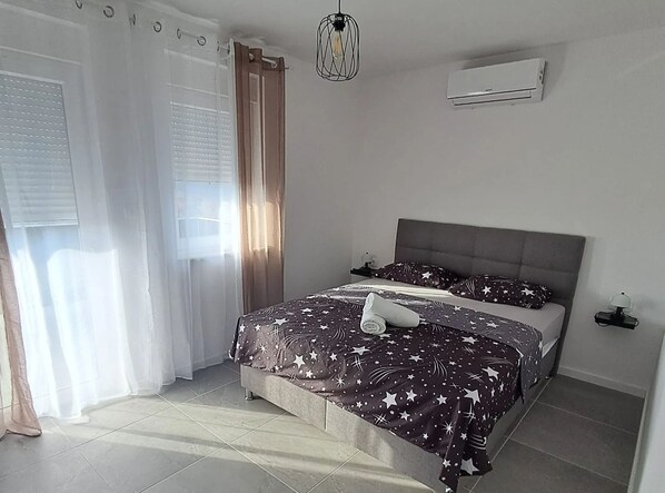 Un dormitorio con cama, cortinas, lámpara colgante, unidad de aire acondicionado y mesita de noche.