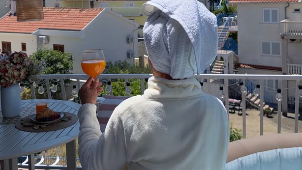 Eine Person hält ein Glas mit Orangensaft, stehend auf einem Balkon mit Pool, umgeben von Gebäuden.