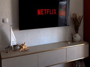 A modern living room with a television displaying 'NETFLIX' in red letters, a small sailboat model, a seashell, and a vase with dried plants on a minimalist console table. - Tina Sea View Retreat (Vodice)