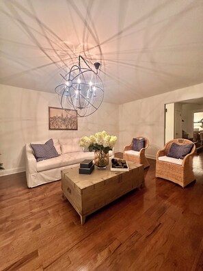 A living room with a white sofa, wicker chairs, a wooden coffee table, and a cityscape painting on the wall. - Normandy H. a charming house in silver spring. 5 min from The Adventist Hospital (Silver Spring)