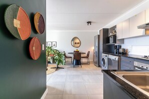 A modern kitchen with a built-in washing machine, a dining area with a round table, and a large mirror on the wall. - Neo-African apartment at the entrance of Durbanville wine route (Cape Town)