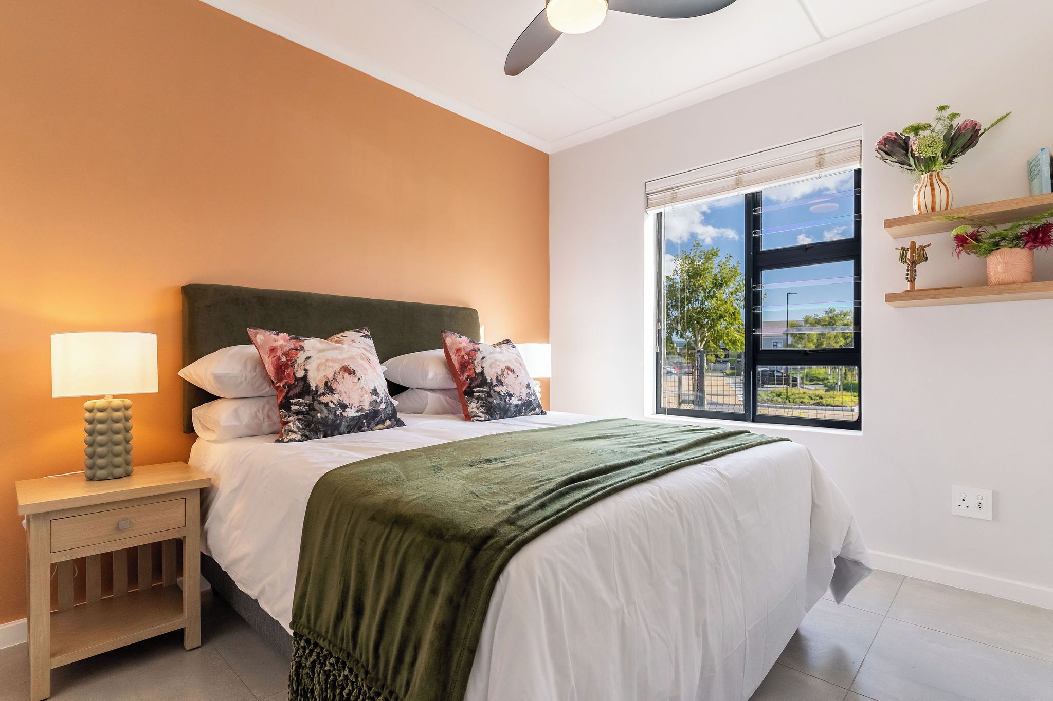 Un dormitorio con una cama, una mesita de noche, una lámpara, un ventilador de techo, una ventana con vistas y un estante con objetos decorativos.